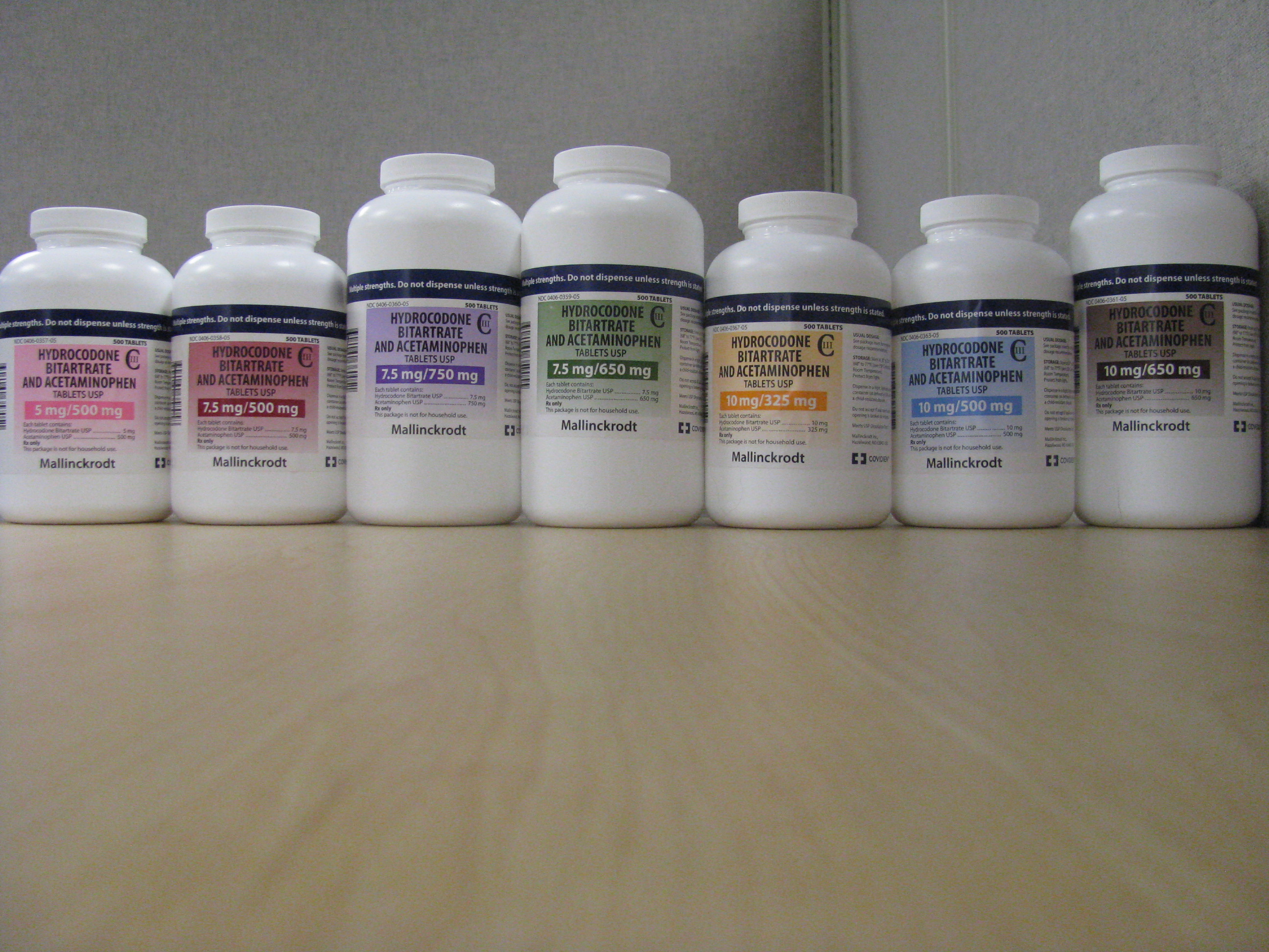 A row of seven white plastic bottles arranged in a line on a wooden surface. The bottles are of different sizes and shapes with different color labels. The labels indicate that the bottles contain hydrocodone bitartrate and acetaminophen with each bottle having a different dosage. The labels also show the Mallinckrodt logo. The background is a plain white wall.