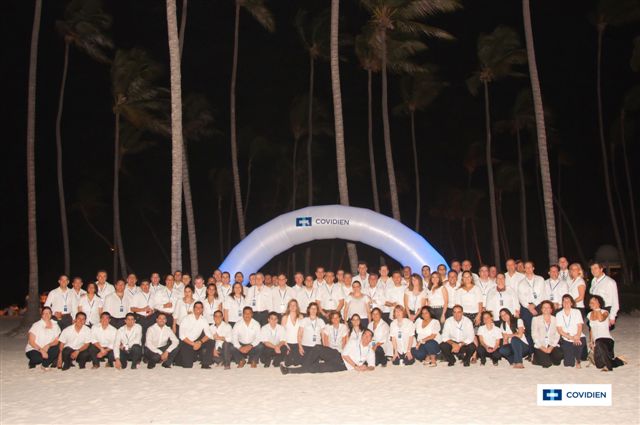 A large group of people posing for a group photo on a beach at night. The group is arranged in a semi-circle with a large blue inflatable arch in the center. The arch has the word "COVIDIEN" written on it in white letters. The people are all wearing white shirts and black pants and they are all smiling and looking at the camera. The beach is lined with palm trees and the sky is dark suggesting that it is nighttime. The overall mood of the photo is celebratory and happy.