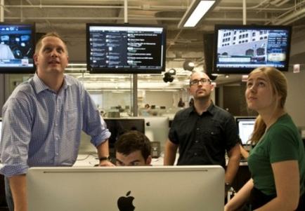 A group of four people in an office setting. There are three people in the foreground two men and two women standing in front of a large computer monitor. The man on the left is wearing a blue checkered shirt and is looking up at the monitor with a serious expression on his face. The woman on the right is sitting at a desk with a laptop in front her. In the background there are multiple computer monitors displaying various images and text. The room appears to be modern and well-lit with fluorescent lights.