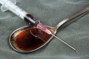 A syringe resting on a spoon that has a dark brown liquid in it likely an opioid or another drug. They are resting on a gray fabric background. The needle is pointing towards the syringe as if it is about to inject it into the spoon.