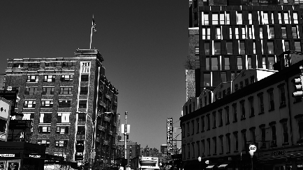 A black and white photograph of a busy city street. The photo is taken from a low angle looking up at a tall building on the left side of the image. The building appears to be a two-story structure with a flat roof and multiple windows. On top of the building there is a flagpole with an American flag flying in the wind. On the right side there are several other tall buildings with balconies and balconies. The street is lined with shops and restaurants and there are traffic lights and street lamps visible. The sky is overcast and the overall mood of the photo is gloomy.