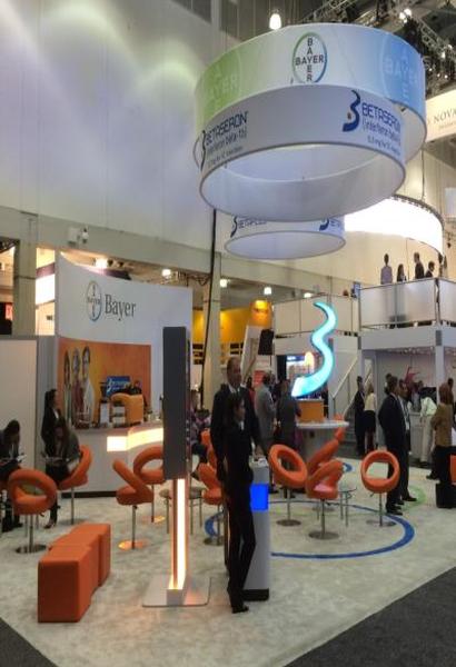 A trade show booth with a large white circular structure hanging from the ceiling. The structure has the Bayer logo and the words "Bayer" written on it. There are several orange and blue chairs and tables arranged around the booth and a few people can be seen walking around the area. The booth appears to be well-lit and has a modern design.