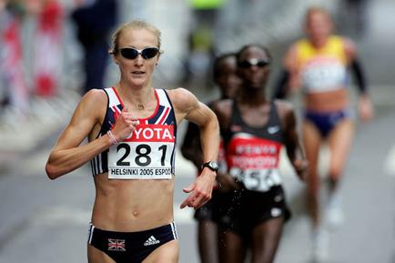 A female athlete running on a road. She is wearing a red and white sports bra with the number 281 on it and black shorts. She has blonde hair and is wearing sunglasses. In the background there are other runners and spectators. The athlete appears to be in the middle of a race as she is running with determination and focus. The image appears to have been taken during the day.