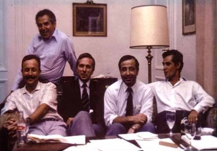A group of five men sitting around a table in a formal setting. The table is covered with papers and a lamp and there are several framed pictures hanging on the wall behind them. The men are all dressed in formal attire with the man on the left wearing a blue shirt and the man in the center wearing a black suit and tie. They are all smiling and appear to be engaged in a conversation. On the right side of the image there is a man standing behind the table looking at the camera. The overall mood of the photo is relaxed and casual.