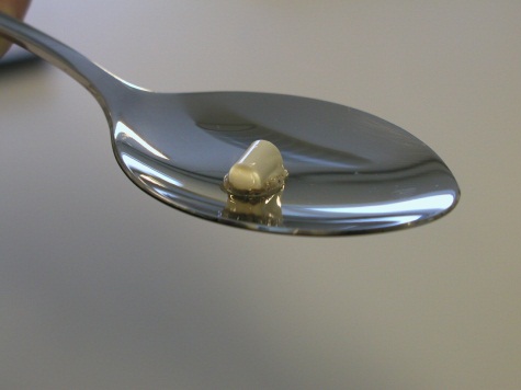 A close-up of a silver spoon with a small amount of white substance on it likely an opioid or another drug. The background is a solid gray color and the focus is on the spoon.