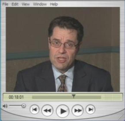 A screenshot of a video player interface. It shows a man in a suit and tie speaking into a microphone. He appears to be in the middle of a conversation. The background is a plain white wall with a blue border. At the top of the screen there is a menu bar with options such as File Edit View Window Help and Help. Below the menu bar there are three buttons - "File" "Edit" "View" and "Window Help". The time "00:18:01" is displayed at the bottom of the image.