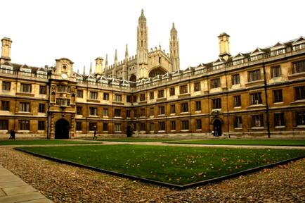 A large historic building with a courtyard in the center. The building is made of stone and has multiple levels with arched windows and balconies. The courtyard is well-maintained with a well-manicured lawn and a stone pathway leading up to the entrance. In the background there is a large cathedral with two tall spires and a clock tower. The sky is overcast and the overall atmosphere of the image is peaceful and serene.