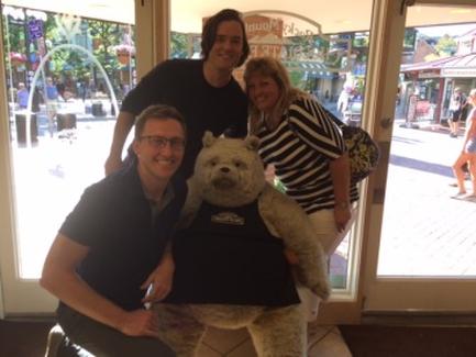 Three people posing with a large teddy bear in front of a store. The person on the left is wearing a black t-shirt and glasses the person in the middle is standing behind the bear and the person at the far right is standing next to the bear. All three people are smiling and appear to be happy. In the background there are shops and people walking on the street.