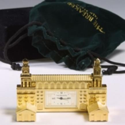 A small gold-colored clock with a green velvet pouch. The clock has a rectangular shape with a white face and black hands. The body of the clock is made up of small intricate details including a clock face with black numbers and hands and two towers on either side. The pouch is made of dark green velvet and has a drawstring closure. The words "The Breakers" are written in gold on the top right corner of the pouch.