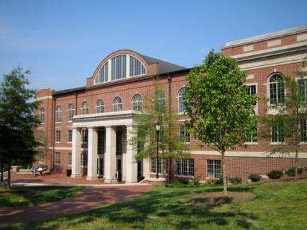 A large brick building with a curved roof and large arched windows. The building is located on a grassy area with trees and shrubs surrounding it. There is a walkway leading up to the entrance of the building and a few people can be seen sitting on benches on the sidewalk. The sky is blue and the weather appears to be sunny and clear.