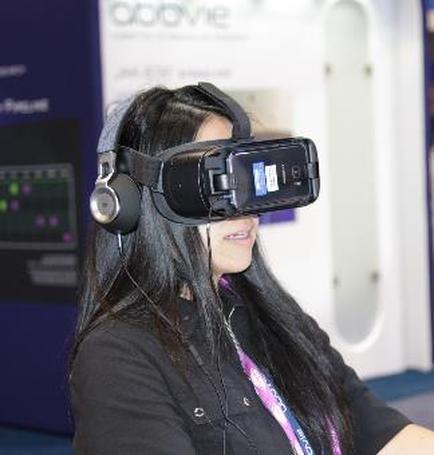 A young woman wearing a virtual reality headset. The headset is black and has a large screen on the front. The woman is wearing a black shirt and has long dark hair. She is looking at the screen with a smile on her face. In the background there is a blue banner with the word "Oddvie" written on it. The image appears to be taken at an exhibition or trade show.