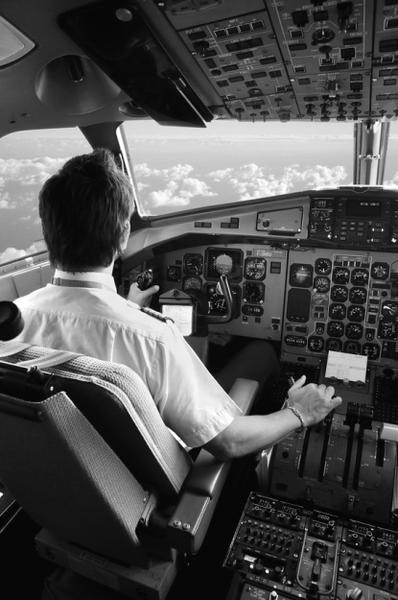 A black and white photograph of a man sitting in the cockpit of an airplane. He is wearing a white shirt and is looking out the window at the clouds outside. The man is holding a control panel in his hands and appears to be operating the instrument panel. The cockpit is filled with various buttons switches and gauges and there is a large window on the right side of the image that offers a view of the sky. The image is taken from a high angle looking down on the man and the clouds.