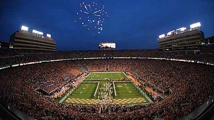 Of a football stadium at night. The stadium is filled with a large crowd of people all of whom appear to be cheering and enjoying the game. The field is covered in green turf and there is a large screen in the center of the field. Above the field there are fireworks exploding in the sky creating a beautiful display of light and color. The sky is dark indicating that it is either night or early morning. The image appears to be taken from the upper level of the stadium looking down on the field and the crowd.