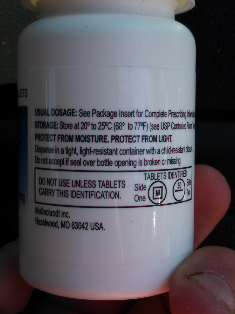 A close-up of the back of a white pill bottle. The label on the bottle is black and white and has text written on it. The text reads "Usual dosage: See Package Insert for Complete Prescription Information. Storage: Store at 20°C to 25°C (68°C) to 77°F (see USP Controlled Room). Protect from moisture. Protect from light. Dispense in a tight light-resistant container with a child-resistant closure. Do not accept if seal over bottle opening is broken or missing. Do NOT USE UNLESS TABLETS carry this identification." It shows an illustration of the tablets which has the letter M on one side and the number 30 on the other. It also has the text "Mallinckrodt Inc." with a corporate address. The background is dark and the bottle appears to be held in a person's hand.
