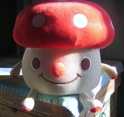 Of a small stuffed toy sitting on top of a wooden box. The toy is white with a red mushroom-shaped cap on its head. The cap has two small white dots on it and the mushroom has a big smile on its face. It has two arms and two legs and it is wearing a blue and white striped shirt. The background is dark and there is a window visible on the right side of the image.