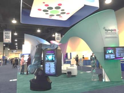 A trade show booth with a large screen on the right side. The booth has a curved design with a green and white color scheme. Above the screen there is a large banner with colorful circles in red green and blue. On the left side of the booth there are several people walking around and a sign that reads "Aisle 300". In the center of the image there appears to be a robotic arm with a screen displaying information about the booth. The floor is covered in a green carpet.