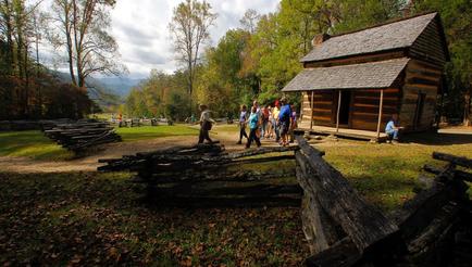 A group of people walking towards a small wooden cabin in a wooded area. The cabin is made of logs and has a sloping roof and a chimney. There is a wooden fence in front of the cabin and a pile of logs on the left side of the image. The ground is covered in fallen leaves and there are trees in the background. The sky is blue and the overall atmosphere is peaceful and serene. The people in the image appear to be tourists visiting the cabin.