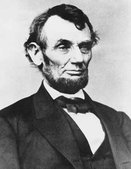 A black and white portrait of Abraham Lincoln the 16th President of the United States. He is shown from the waist up looking directly at the camera with a serious expression on his face. He has a full beard and mustache and his hair is neatly combed back. He appears to be wearing a suit and tie with a collared shirt underneath. The background is plain and white making the portrait the focal point of the image.