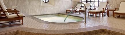 An indoor hot tub in a luxurious spa-like setting. The hot tub is in the center of the image and is surrounded by a tiled floor. There are several wooden lounge chairs with white cushions on either side of the tub arranged in a semi-circle around it. The chairs have a modern design with a curved backrest and armrests. The walls are covered in white marble tiles and there is a large window on the right side allowing natural light to enter the space. A metal railing runs along the edge of the hot tub providing access to the water. The overall atmosphere of the space is peaceful and relaxing.