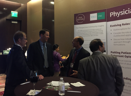 A group of four men gathered around a table in a conference room. They are engaged in a conversation and appear to be engaged in some kind of activity. The table is covered with a white tablecloth and there is a glass of red wine on the table. On the right side of the image there are two banners with the words "Physician" and "Evolving Roles" written on them. The banner on the left side has a list of different roles and responsibilities of the participants. The background shows a wall with a sign that reads "CNS" and a door. The men are dressed in formal business attire and are dressed casually.