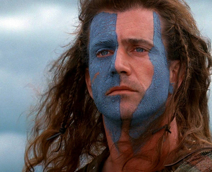 A close-up portrait of a man's face and upper body. He has long curly hair that is styled in loose waves and falls over his shoulders. His face is painted in a blue and white striped pattern with the blue being the dominant color. The man's eyes are dark and his eyebrows are furrowed. He is looking off to the side with a serious expression on his face. The background is blurred but it appears to be a cloudy sky. The overall mood of the image is somber and contemplative.