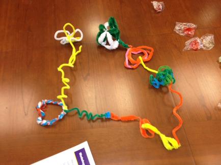 A wooden table with a colorful string art project on it. The string art is made up of different colors and shapes including yellow green orange blue and red. There are also two small white flowers made of pipe cleaners attached to the string. On the right side of the table there are several small packets of candy and on the left side there is a piece of paper with a purple label. The background is blurred but it appears to be a classroom or classroom setting.