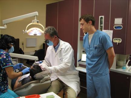 A dental clinic with three people in it. On the left side of the image there is a dentist wearing a white coat and a face mask sitting in a dental chair with his mouth open. He is holding a dental instrument in his hand and appears to be examining a patient's teeth. Next to him there are two dental professionals one wearing a blue scrub suit and the other wearing a pink shirt and blue gloves. The dentist is standing next to the patient who is sitting in the chair and is looking at him attentively. The patient is lying on the dentist's chair with their eyes closed and their mouth slightly open. There is a dental lamp above them and a desk with various dental tools and equipment in the background. The room is well-lit with natural light coming in from the window.