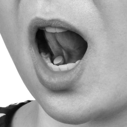 A close-up of a person's mouth. The person appears to be in their late twenties or early thirties. Their mouth is open wide with their tongue sticking out and their teeth visible. The image is in black and white and the person is wearing a polka dot shirt. The background is plain white.