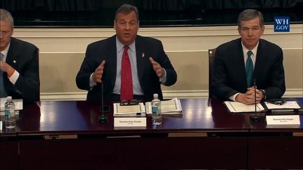Three men sitting at a long wooden table in a meeting room. The man in the center is speaking into a microphone and appears to be giving a speech. He is wearing a suit and tie and has a serious expression on his face. On the left side of the table there is another man sitting next to him also speaking into the microphone. The other two men are listening attentively. All three men are wearing suits and appear to be in a formal setting. The table is covered with papers water bottles and nameplates. The background of the image is a plain white wall with a black curtain.