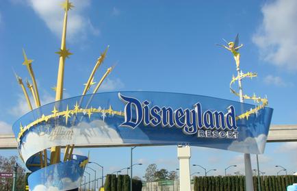 Of the entrance of Disneyland Resort. The entrance is a large blue sign with the words "Disneyland Resort" written in white capital letters. Above the sign there is a golden archway with three golden stars on either side. On top of the archway there are two golden statues of fairies one on each side. The fairies are wearing green dresses and have wings. The sky is blue with white clouds and there are trees and bushes in the background.