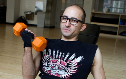 A man sitting on a chair in a gym holding a pair of orange dumbbells in his hands. He is wearing a black sleeveless t-shirt with a graphic design on it and black gloves. He has a bald head and glasses and is looking directly at the camera with a serious expression on his face. The background shows a wooden floor and a bookshelf with various exercise equipment.