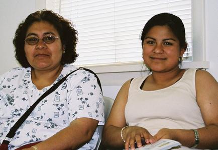 Two women sitting side by side on a couch. The woman on the left is wearing a white blouse with a blue floral pattern and glasses. She has shoulder-length dark hair and is looking directly at the camera with a serious expression. The other woman is sitting next to her wearing a light-colored tank top and has dark hair. She is holding a piece of paper in her hand and is smiling at the viewer. The background shows a window with white blinds.