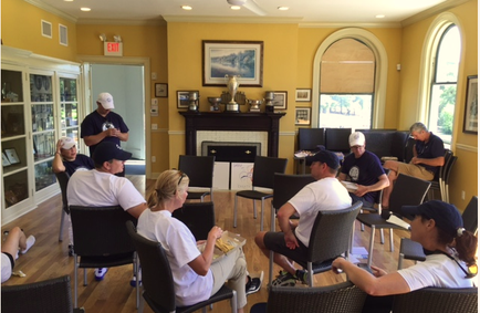 A group of people sitting in a living room with a fireplace in the background. The room has yellow walls and a large arched window on the right side. There are several trophies on the fireplace mantel and a painting hanging on the wall. The people in the room are of different ages and genders and they are all wearing casual clothing and baseball caps. They are engaged in a conversation and appear to be engaged in some kind of activity. There is an exit sign on the left side of the room and a bookshelf on the far wall.