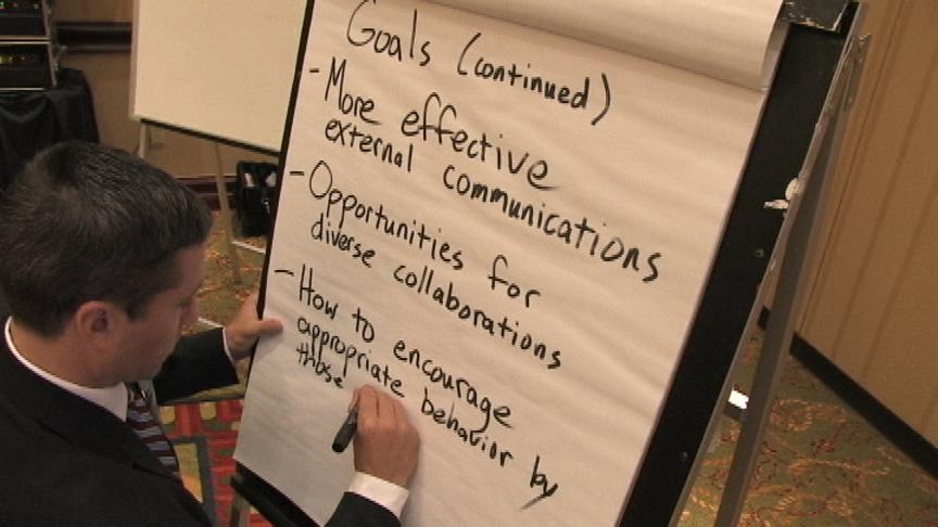 A man in a suit writing on a large easel pad. The pad has a list of goals written on it in black marker. It reads: <br /><br />Goals (Continued)<br />- More effective external communications<br />- Opportunities for diverse collaborations<br />- How to encourage appropriate behavior by those<br /><br />The man is holding a marker in his right hand and and continues to write the final bullet point. He seems to be focused on the task at hand. The background of the image is a conference room with a carpeted floor.