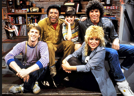 A group of five young people posing for a photo in a room with a brick wall and a bookshelf in the background. There are six people in the photo three men and three women all of whom are smiling and looking at the camera. The man on the left is wearing a striped shirt and suspenders the man in the middle is wearing an orange jumpsuit and the woman on the right is sitting on the floor with her legs crossed. All of them are wearing casual clothes and appear to be happy and relaxed.