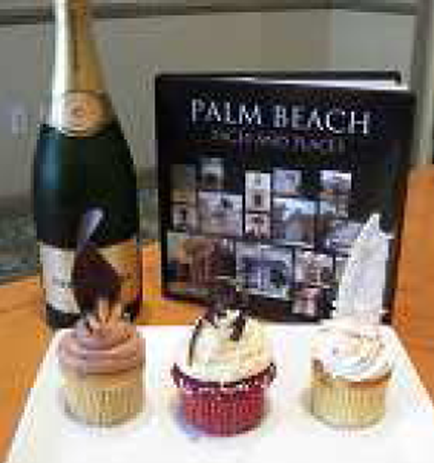A table with a bottle of champagne and three cupcakes on it. The bottle is on the left side of the table and the cupcakes are on the right side. The cupcakes have white frosting and are topped with chocolate shavings. In the background there is a black book titled "Palm Beach" with a collage of photos on the cover. The table appears to be in a kitchen or dining area.