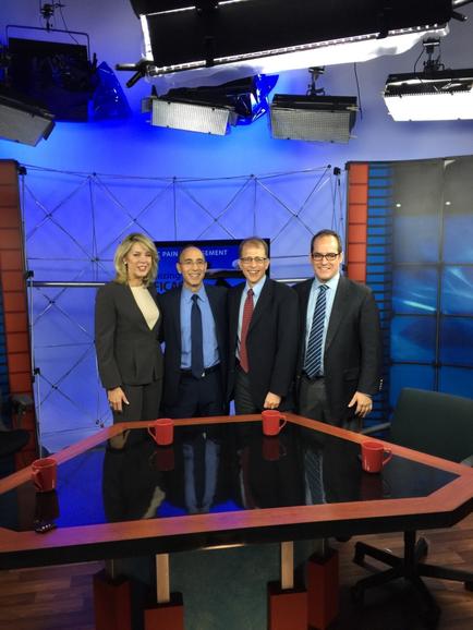 A group of five people standing in front of a news studio set. There is a large table in the center of the room with red coffee mugs on it. The people are all dressed in formal business attire and are smiling at the camera. Behind them there is a blue wall with a large screen and several spotlights hanging from the ceiling. On the left side of the image there are two chairs and a desk. The overall atmosphere of the studio appears to be professional and modern.