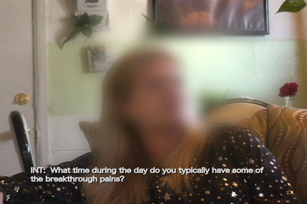 A person sitting on a couch in a living room. The person is blurred but their face is not visible. They are wearing a black top with white stars on it. The background shows a green wall with a picture hanging on it and a vase with red flowers. There is also a door visible in the background. The text on the image reads "INT: What time during the day do you typically have some of the breakthrough pains?"