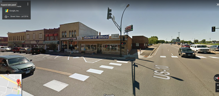 A street view of a small town with a few buildings on the left side of the image. On the right side there is a building with a sign that reads "temper Drug" and a store front with a blue awning. The building appears to be a convenience store or a drugstore. There are several cars parked on the street in front of the building and a traffic light on the corner. The sky is blue and there are a few trees in the background. The street is empty with no cars or people visible.