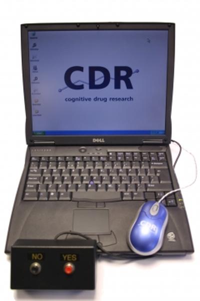A Dell laptop with a blue screen displaying the logo of Cognitive Drug Research (CDR) on it. The laptop is placed on a white surface and there is a blue computer mouse next to it. On the left side of the laptop there is an electronic device with two buttons - "NO" and "YES" - which is likely used to control the laptop's settings. The device appears to be connected to the laptop via a cable.