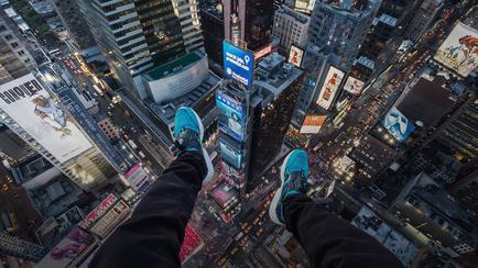 A pair of feet standing on top of a skyscraper in New York City. The feet are wearing black pants and blue sneakers. The skyscraper is surrounded by other tall buildings and skyscrapers and there are advertisements and billboards visible on the buildings. The image is taken from a high vantage point looking down on the city below. The sky is dark indicating that it is nighttime.