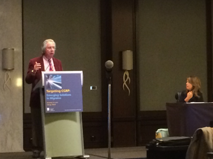 A man standing at a podium in a conference room. He is wearing a red suit and tie and appears to be giving a presentation. The podium has a blue banner with the words "Targeting CRP" written on it. There is a microphone on the right side of the podium and a woman sitting at a table with a microphone in front of her. The room has a beige wall and a carpeted floor.
