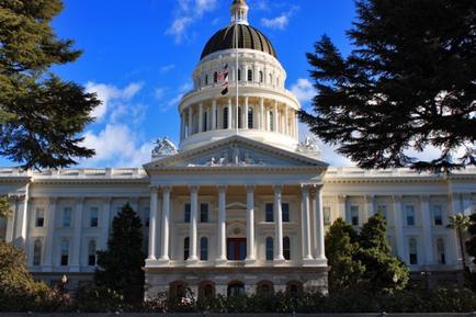 Of the California State Capitol building in Sacramento California. The building is a large white neoclassical structure with a dome on top and a flagpole with the American flag flying in front of it. The dome is decorated with intricate carvings and columns and the building is surrounded by trees and greenery. The sky is blue and there are a few clouds in the background. The image is taken from a distance looking up at the building from across the street.