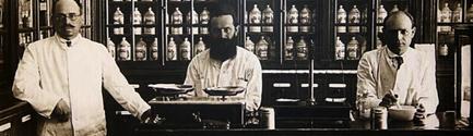 A black and white photograph of three men in a laboratory. They are all wearing lab coats and appear to be in their late twenties or early thirties. The man on the left is standing with his hands on his hips while the man in the middle is sitting at a desk with a plate of food in front of him. All three men have beards and are looking at the camera. Behind them there are shelves filled with various bottles and jars. The photograph appears to be from the early 20th century.