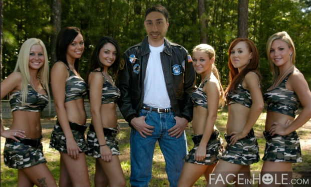 A group of six young women and a man posing together in a wooded area. The man is standing in the center of the group wearing a black leather jacket with patches on it and a white t-shirt. He has a mustache and is smiling at the camera. The women are all wearing matching camouflage outfits with one woman on the left wearing a green tank top and shorts another wearing a white tank top and the third wearing a blue tank top. They are all standing close together with their arms around each other's waist. The background is filled with trees and greenery creating a peaceful and natural setting.