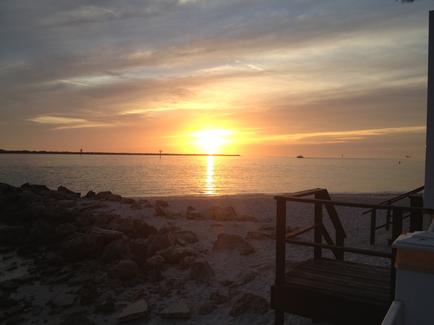 A beautiful sunset over the ocean. The sky is filled with orange and yellow hues with the sun partially hidden behind the clouds. The sun is setting over the horizon casting a warm glow over the water. The water is calm and reflects the colors of the sky. On the right side of the image there is a wooden deck with a railing and a table and chairs overlooking the beach. The beach is covered in rocks and pebbles and there are a few palm trees in the background. The overall mood of the photo is peaceful and serene.