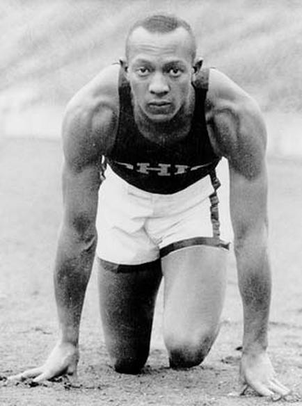 A black and white photograph of a man in a track and field setting. He is wearing a sleeveless tank top and shorts and his body is in a crouching position with his hands on the ground. He appears to be in the middle of a race or competition as he is looking directly at the camera with a serious expression on his face. The background is blurred but it seems to be a grassy field with trees in the distance. The photograph is likely from the early 20th century.