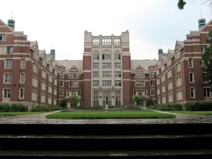 A large brick building with multiple floors and a tall tower in the center. The building has multiple windows and is surrounded by a well-manicured lawn with a fountain in the middle. The sky is overcast and there are trees and bushes on either side of the building. In the foreground there are steps leading up to the entrance. The overall atmosphere of the image is peaceful and serene.