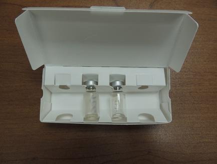 A white plastic box with a lid on top. Inside the box there are two small glass vials with silver caps. The vials appear to be empty and are placed on a wooden surface. The box has two compartments on the sides one on each side for storing the vials. The lid of the box is open revealing the contents inside.