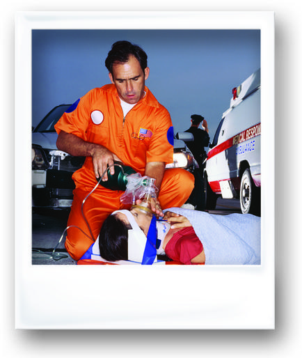 A man in an orange jumpsuit with an American flag on his chest kneeling down next to a woman lying on a stretcher. The man is holding an oxygen mask and appears to be performing an emergency medical procedure on the woman's face. The woman is wearing a white hospital gown and is lying on her back with her eyes closed. In the background there is a white ambulance and a police officer. The image appears to have been taken at night as the sky is dark and the ambulance is parked on the side of the road.